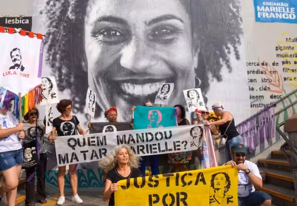 People holding banners and posters demanding justice for Marielle Franco and asking who is behind her assassination, in front of a mural photo of the Brazilian LGBTQ activist and politician. Photo: Scuzinska/Alamy Stock Photo/File photo.