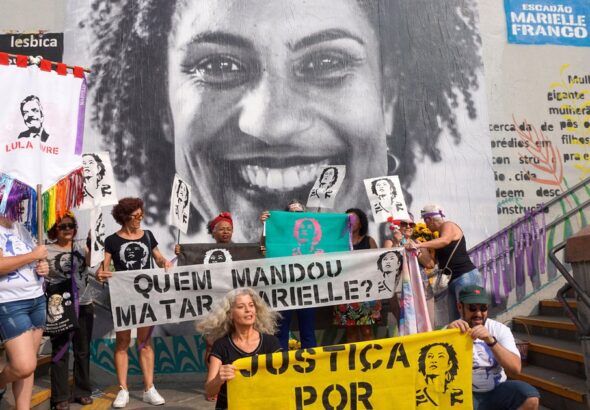 People holding banners and posters demanding justice for Marielle Franco and asking who is behind her assassination, in front of a mural photo of the Brazilian LGBTQ activist and politician. Photo: Scuzinska/Alamy Stock Photo/File photo.