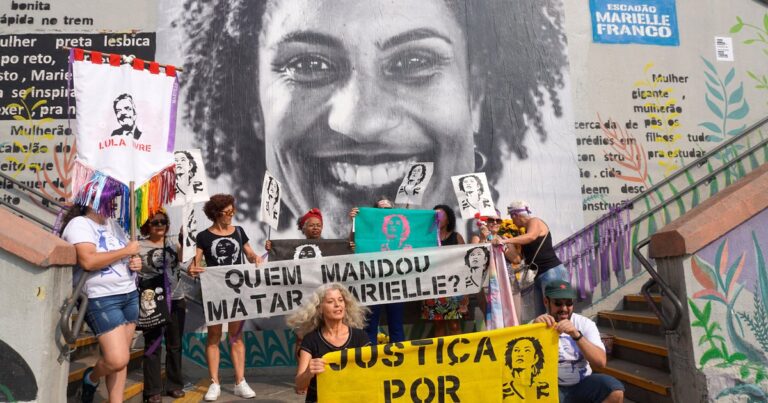 People holding banners and posters demanding justice for Marielle Franco and asking who is behind her assassination, in front of a mural photo of the Brazilian LGBTQ activist and politician. Photo: Scuzinska/Alamy Stock Photo/File photo.