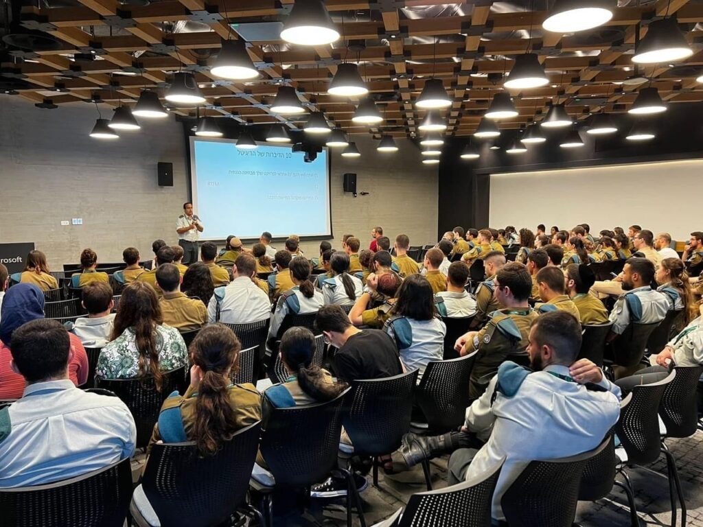 A packed room of Israel Defense Forces (IDF) soldiers attending a conference at a Microsoft Israel R&D center. The photo was publicly shared by the Lotem technology unit of the IDF’s Computer Services Directorate on August 2, 2023 and shows a conference led by the unit’s Shahar component the previous day, with roughly 150 soldiers in attendance. Microsoft’s logo can be partially seen on the center of the left side of the image and the title of the displayed slide translates from Hebrew to “the 10 commandments of the digital.” The second such commandment can be seen to be “RTFM,” meaning: “Read The Fucking Manual.”