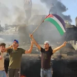 Palestinian young men hold a Palestinian flag in celebration around a destroyed Israeli tank in Gaza City. Photo: Hani Alshaer/Anadolu Agency.
