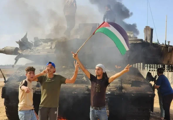 Palestinian young men hold a Palestinian flag in celebration around a destroyed Israeli tank in Gaza City. Photo: Hani Alshaer/Anadolu Agency.