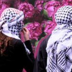 Two people wearing keffiyehs stand facing a wall covered in flowers. Photo: Institute for Palestine Studies.
