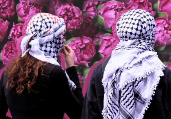 Two people wearing keffiyehs stand facing a wall covered in flowers. Photo: Institute for Palestine Studies.