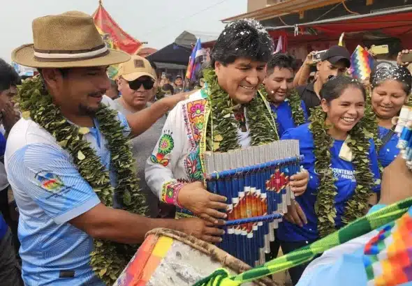 Evo Morales holding a panpipe and wearing a garland as he stands among a crowd. Photo: Telegram/Sputnik Mundo.