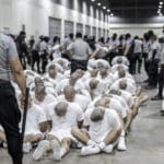 Inmates of the Salvadoran mega-prison CECOT sit on the floor while security personnel stand guard over them. Photo: France24.