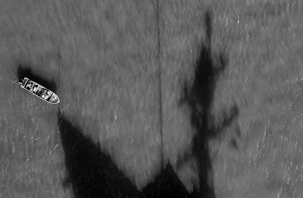 Black-and-white aerial photograph showing a small boat on the water and a large shadow of a US warship. Photo: Matias Delacroix/Associated Press.