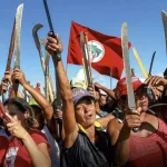 Women members of Brazil’s Landless Rural Workers’ Movement (MST) hold their scythes and machetes aloft. Photo: Brasil Paralelo.