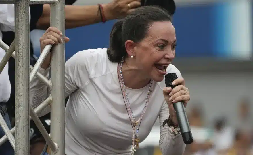 Venezuelan opposition leader María Corina Machado speaks into a microphone at a public event. She is wearing a white shirt and is holding onto a metal railing. Photo: CODEPINK.