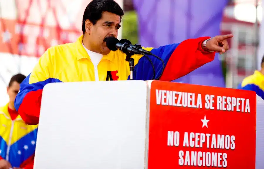 The image shows Venezuelan President NicolĂĄs Maduro at a political rally. He is standing behind a podium with signs that read "Venezuela Se Respeta" and "No Aceptamos Sanciones." These Spanish phrases translate to "Venezuela is respected" and "We do not accept sanctions". Photo: Venezuela Analysis.