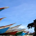The image captures a moment on a Venezuelan beach, featuring several small fishing boats drawn up on the sand and a fisherman carrying an outboard motor over his shoulder. Photo: Pixabay/file photo.
