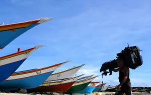 The image captures a moment on a Venezuelan beach, featuring several small fishing boats drawn up on the sand and a fisherman carrying an outboard motor over his shoulder. Photo: Pixabay/file photo.