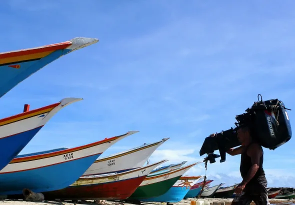 The image captures a moment on a Venezuelan beach, featuring several small fishing boats drawn up on the sand and a fisherman carrying an outboard motor over his shoulder. Photo: Pixabay/file photo.