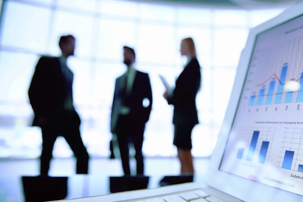 Silhouetted business professionals stand in a modern office as a laptop in the foreground displays financial charts and data analytics. Photo: Rawpixel/Getty Images.