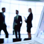 Silhouetted business professionals stand in a modern office as a laptop in the foreground displays financial charts and data analytics. Photo: Rawpixel/Getty Images.