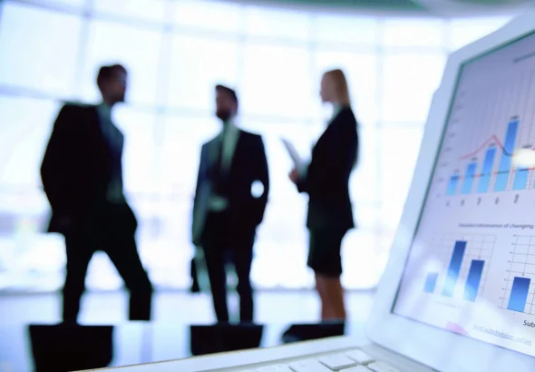 Silhouetted business professionals stand in a modern office as a laptop in the foreground displays financial charts and data analytics. Photo: Rawpixel/Getty Images.