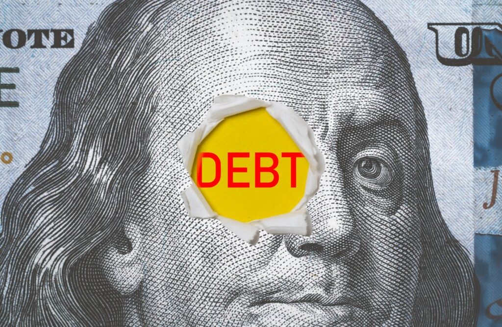 The word "DEBT" written in red on a US dollar bill. Photo: Shutterstock.