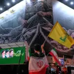 An Iranian demonstrator waves a flag of Lebanon's militant Hezbollah group under an anti US billboard depiciting the American aircrafts into the Iranian armed forces fishing net, during a gathering in Tehran, Iran, May 4, 2026. Photo: AP.