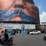 Cars and motorcycles drive by a billboard with graphic showing Strait of Hormuz and the sewn lips of US President Donald Trump in a square in downtown Tehran on May 2, 2026. Photo: PressTV.