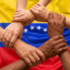 Several hands held in unison, forming a circle in front of the Venezuelan flag. Photo: AI-generated image.