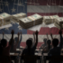 This image depicts students in a classroom raising their hands beneath stacks of money suspended like marionettes, with a U.S. flag in the background. It symbolically suggests the influence of money over education and public institutions. Photo: MintPress News.