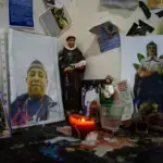 A small domestic altar or shrine featuring religious statues, framed portraits, and devotional items arranged on a dark wooden surface with a burning red candle. Photo: Center for Economic and Policy Research.