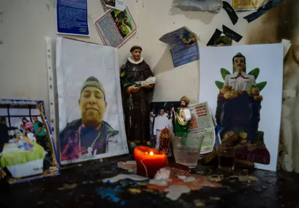A small domestic altar or shrine featuring religious statues, framed portraits, and devotional items arranged on a dark wooden surface with a burning red candle. Photo: Center for Economic and Policy Research.